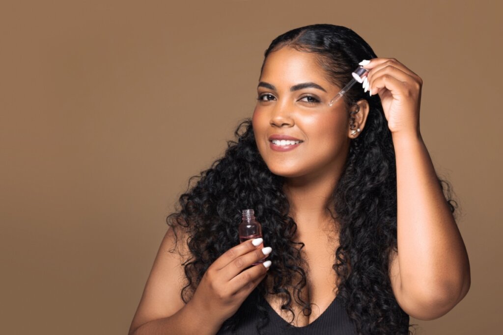 Mulher de cabelos cacheados aplicando produto facial com conta-gotas, segurando um frasco pequeno