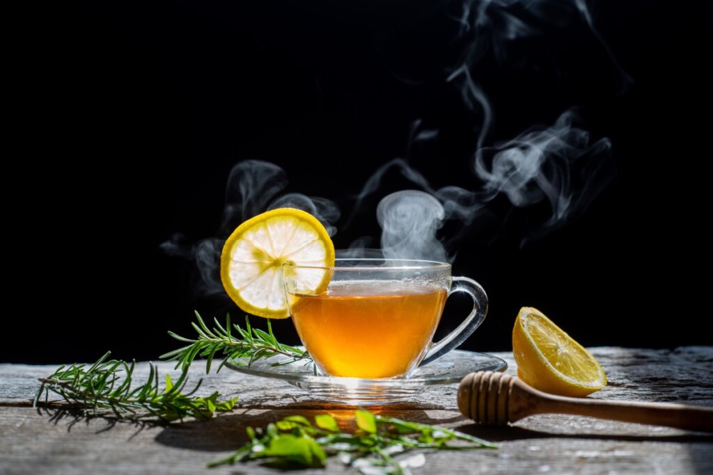 Xícara de vidro com chá quente sobre superfície de madeira, acompanhada por fatia de limão, ramo de alecrim e um pegador de mel de madeira ao lado