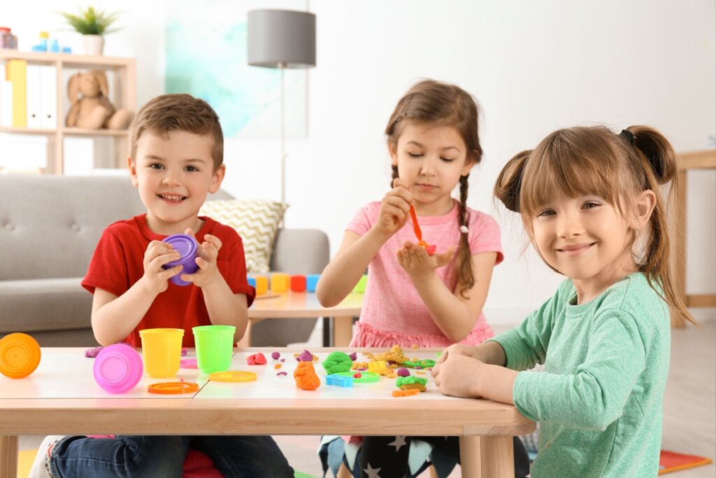 um menino e duas meninas pequenos sentados no chão em volta de mesa baixa e brincando com massinha colorida