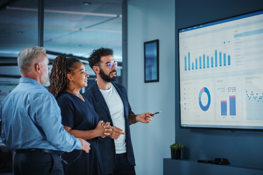 Três pessoas com roupas sociais em tons de azul, analisando dados em telão no escritório