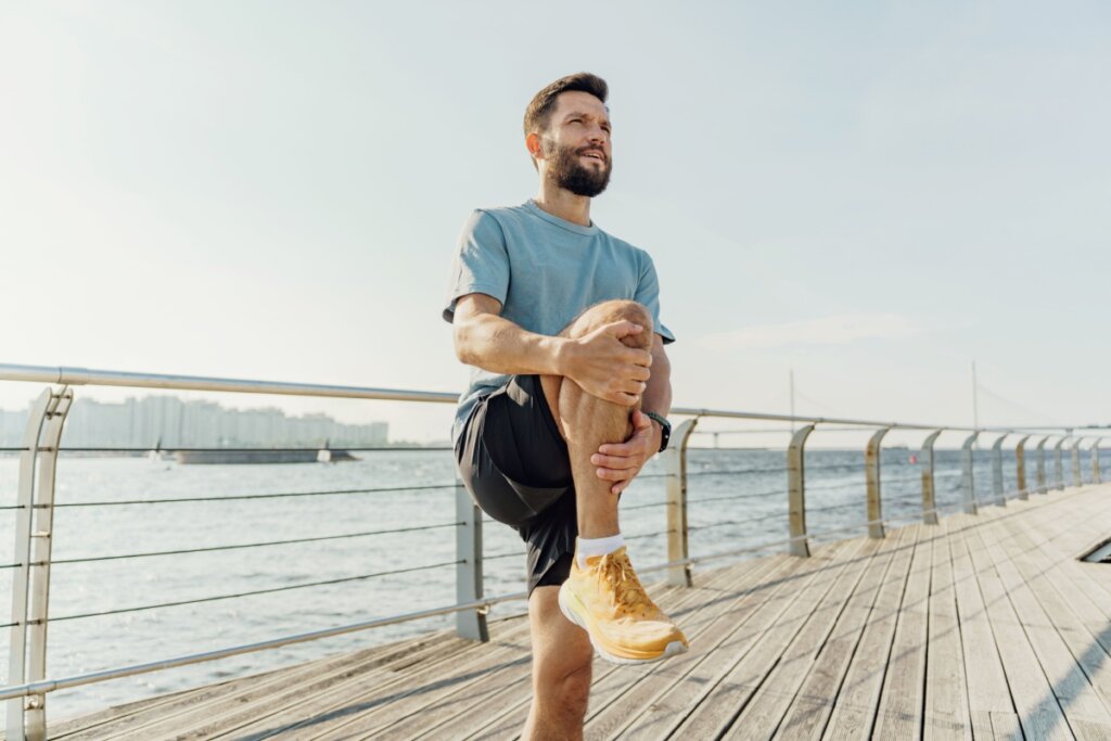Homem com cabelo curto, barba, usando camiseta azul, bermuda preta e tênis amarelo com meia branca alongando ao ar livre