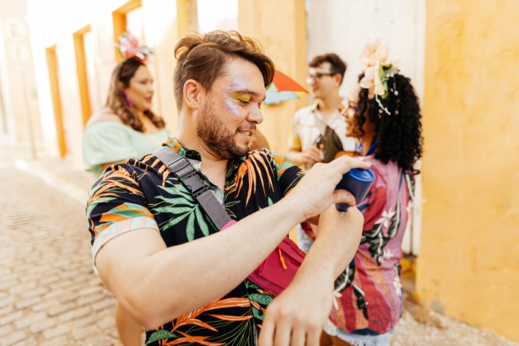 Homem com cabelo curto, camiseta preta estampada passando protetor solar no braço em bloco de carnaval
