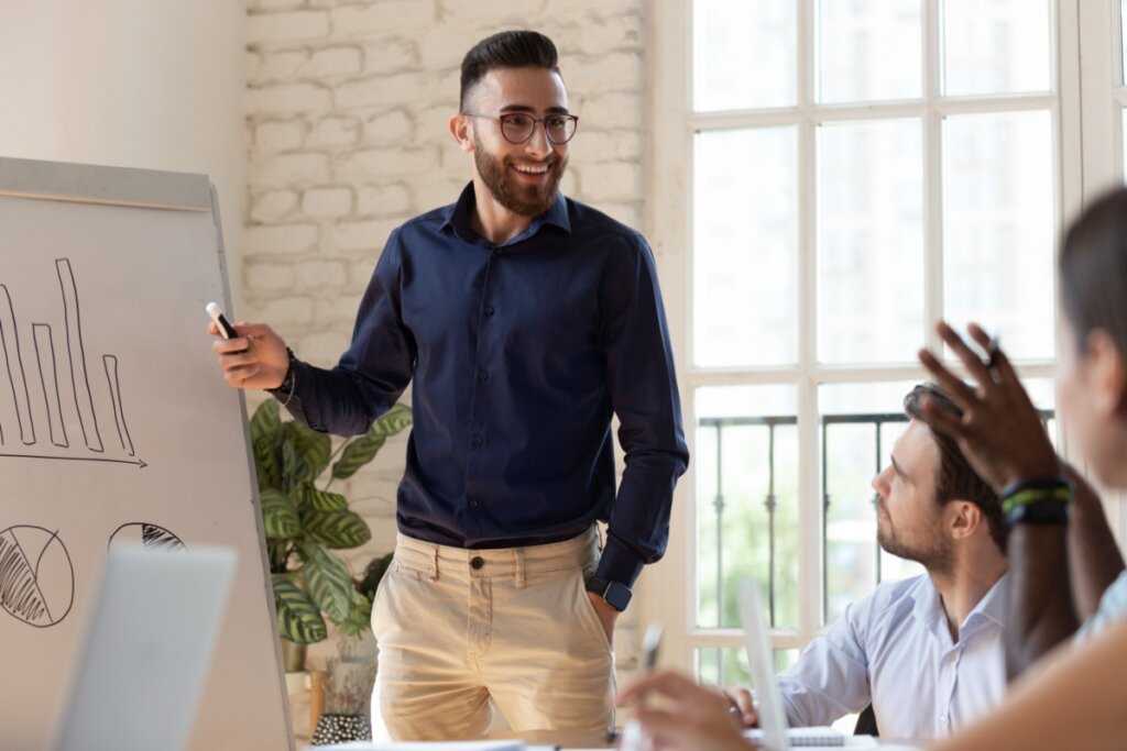 Homem com cabelo curto, barba e usando óculos preto, camisa social azul-escura e calça bege mostrando dados em quadro em reunião de equipe 
