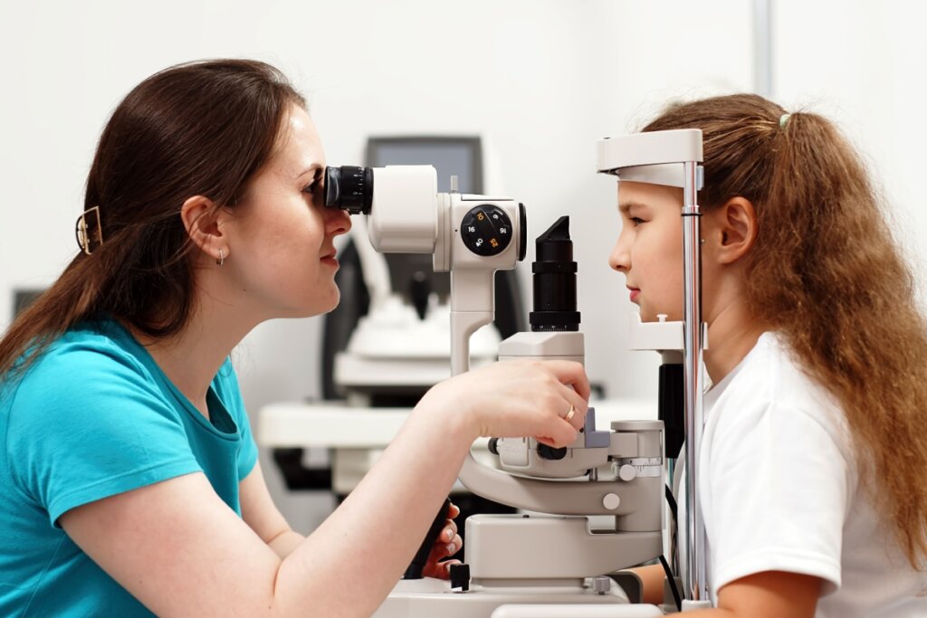 Médica com o cabelo liso, preso parcialmente, usando camiseta verde fazendo exame de vista em criança com o cabelo amarrado, usando camiseta branca com rosto em aparelho 