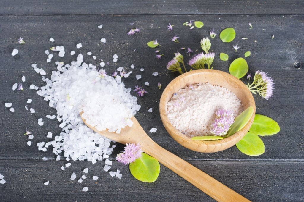 Sal grosso em uma cumbuca ao lado de flores e em uma colher em cima de uma bancada cinza
