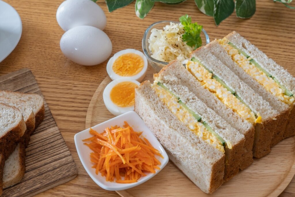 Sanduíche de ovo com pepino servido em tábua de madeira em cima de mesa de madeira, com ovos ao lado e tigela com cenoura ralada