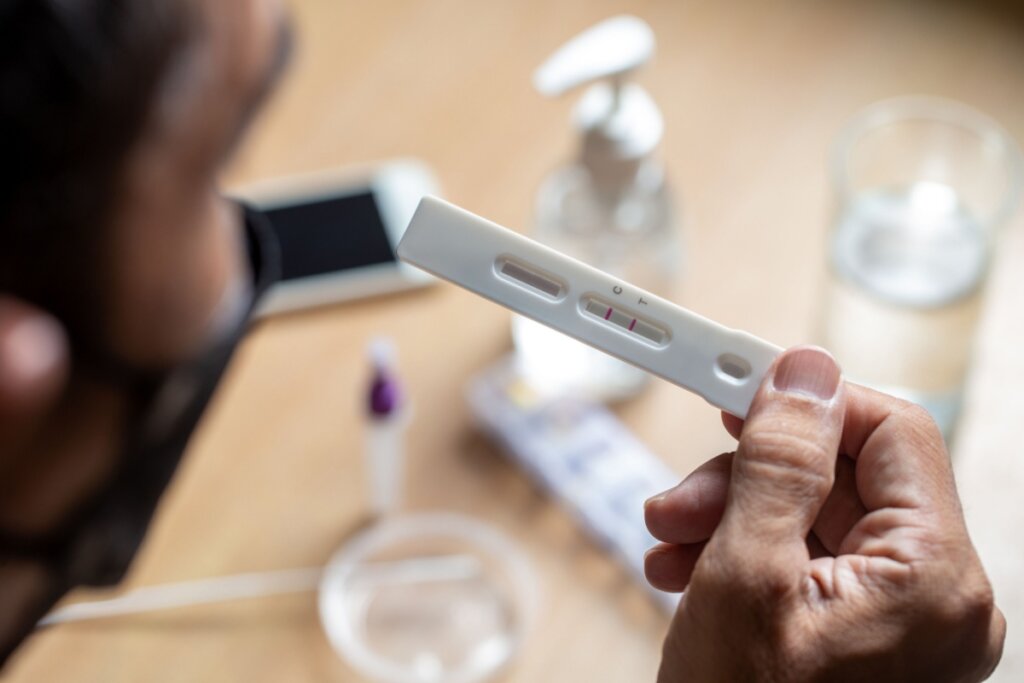 Mão segurando um teste rápido. Ao fundo, mesa de madeira com objetos brancos desfocados