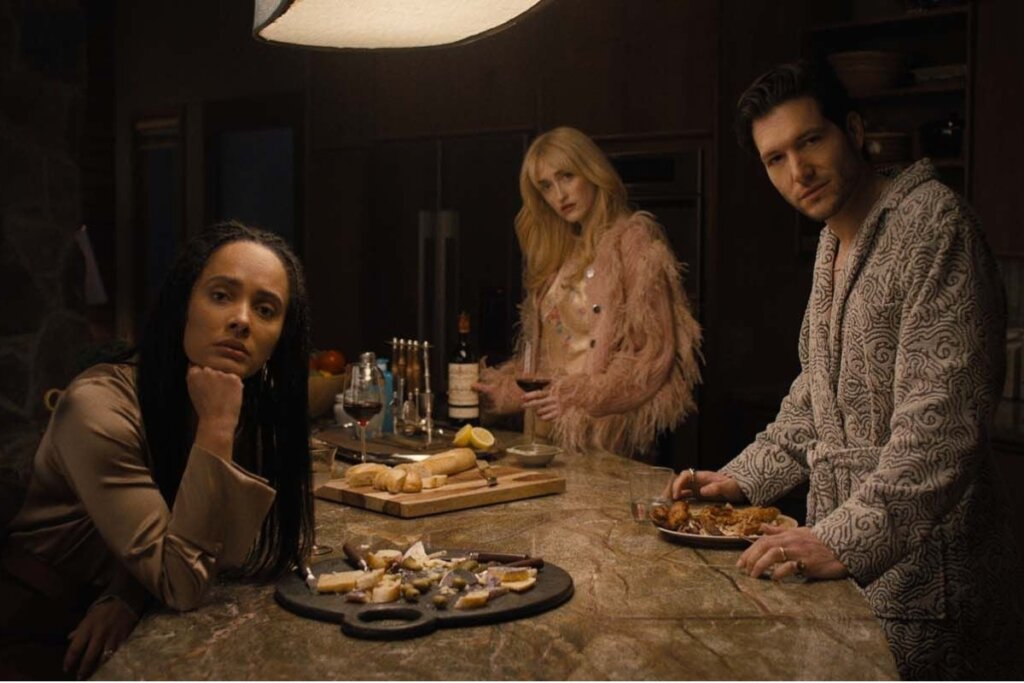 Cena da série "Algo Horrível Vai Acontecer" em uma cozinha de luxo com iluminação baixa e dramática. Três personagens aparecem com expressões sérias e tensas: uma mulher negra à esquerda apoiando o rosto na mão, uma mulher loira ao fundo segurando uma taça de vinho e um homem à direita vestindo um roupão estampado. Sobre a bancada de mármore, há pães, queijos e pratos com comida.