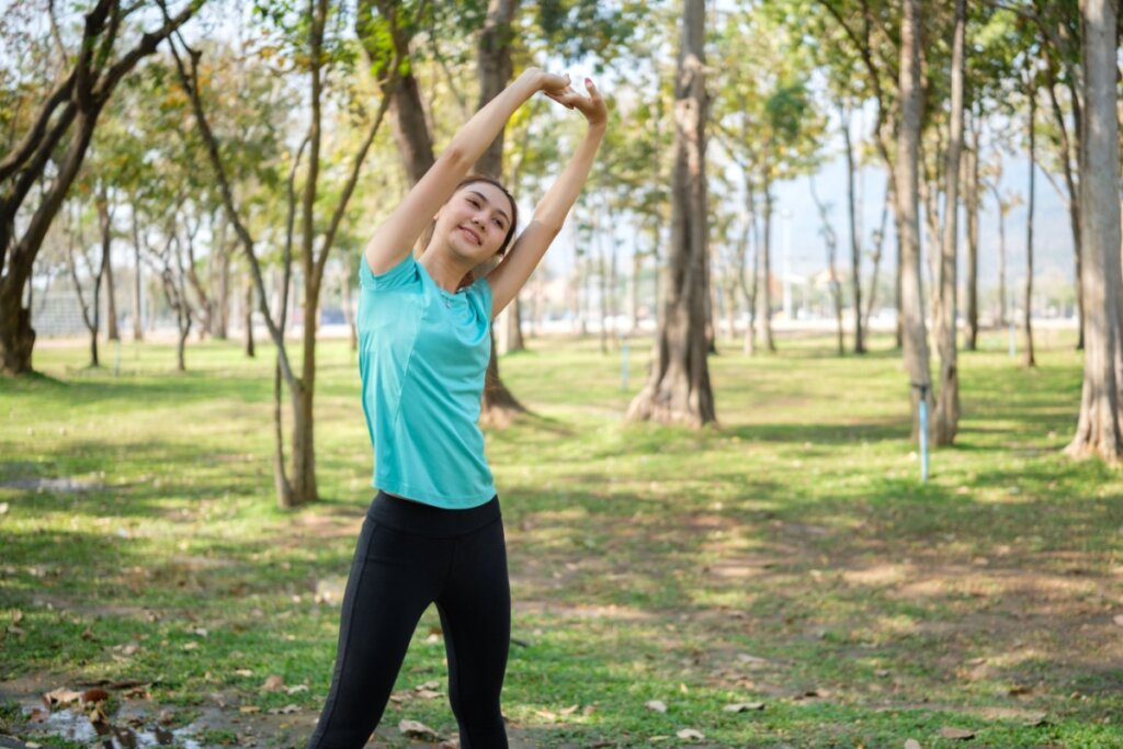 Mulher, usando blusa verde e calça preta, em um parque alongando os braços 