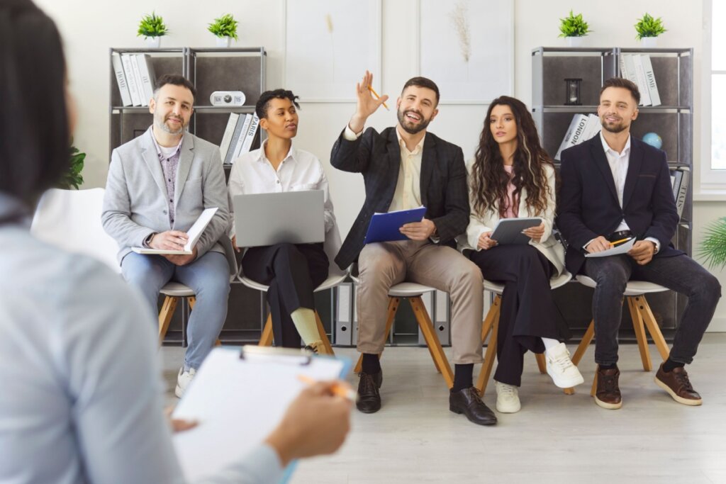 Em uma sala de treinamento, uma palestrante conduz uma sessão de perguntas e respostas. Cinco participantes estão sentados em linha; no centro, um homem levanta a mão com uma caneta para tirar uma dúvida. Os demais colegas acompanham a explicação com materiais de anotação, tablets e laptops.