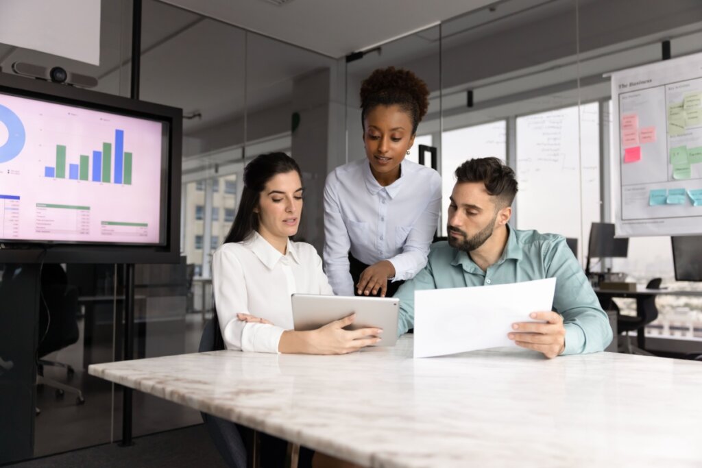 Fotografia de três profissionais — duas mulheres e um homem — em um escritório moderno, colaborando em torno de uma mesa de mármore. À esquerda, uma mulher branca de cabelos escuros segura um tablet; ao centro, uma mulher negra de cabelos presos observa o dispositivo; à direita, um homem branco de barba analisa um documento impresso. Ao fundo, uma tela exibe gráficos coloridos e uma parede de vidro contém anotações, sinalizando um ambiente de trabalho analítico e diversificado.