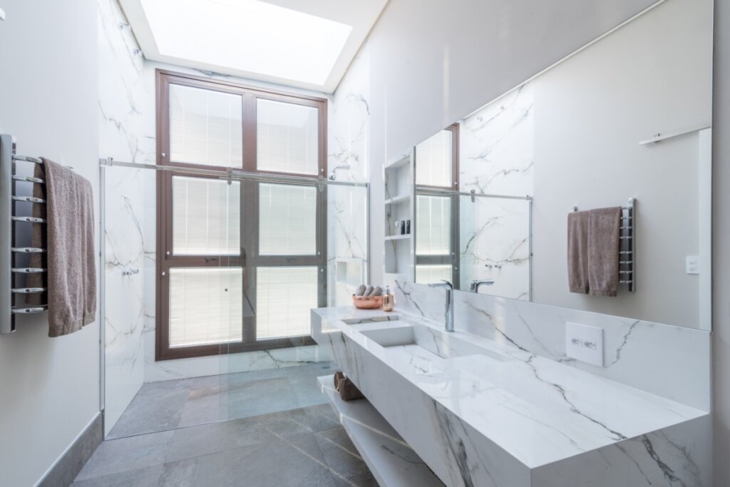 Fotografia de um banheiro contemporâneo com foco na iluminação zenital. O teto possui uma abertura retangular protegida por vidro que permite a entrada abundante de luz natural, iluminando diretamente a área do box. O revestimento das paredes é em tons de cinza claro com textura de cimento queimado. O espaço inclui um chuveiro de teto cromado, um nicho embutido na parede para produtos de higiene e uma bancada de madeira com uma cuba de apoio branca. O ambiente transmite uma estética spa, minimalista e relaxante.