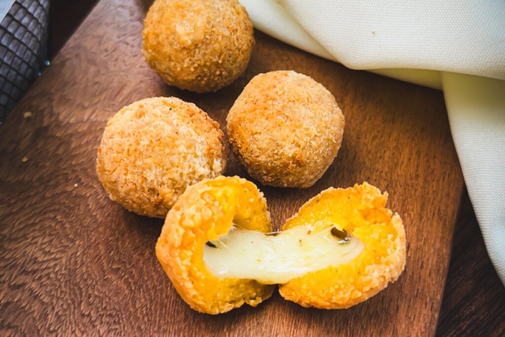 Uma tábua de madeira rústica com quatro bolinhas de queijo fritas e douradas. Uma das bolinhas está partida ao meio em primeiro plano, revelando o recheio de queijo derretido e elástico. Os salgadinhos possuem uma casca crocante e bem empanada. Ao fundo, um guardanapo de tecido claro e dobrado completa a composição, com iluminação quente que destaca a textura crocante do empanado.