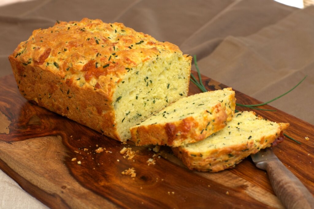 Bolo de fubá com queijo e cebolinha, em cima de uma tábua de madeira, com duas fatias cortadas 