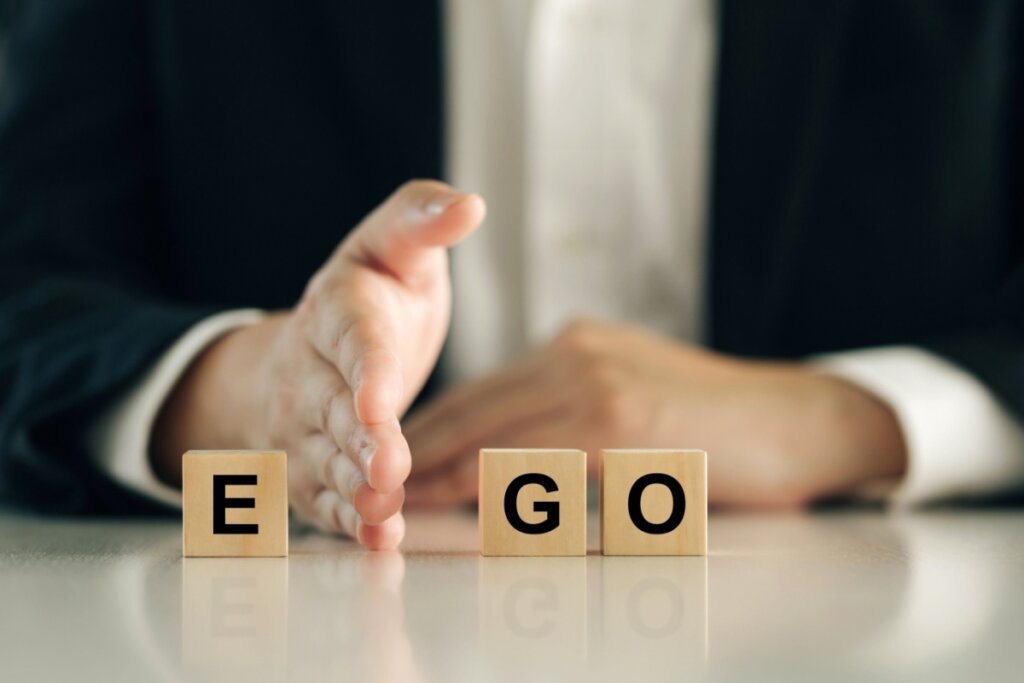 Close das mãos de uma pessoa vestindo paletó escuro, posicionadas atrás de três blocos de madeira que formam a palavra "EGO". Uma das mãos está em pé, como uma barreira, separando a letra "E" das letras "G" e "O". A imagem utiliza um fundo desfocado e tons sóbrios, sugerindo o conceito de controle ou distanciamento do ego.
