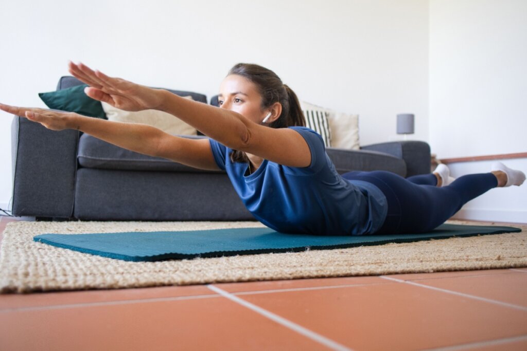 Mulher, usando roupa azul, em uma sala de estar fazendo elevação de braço e perna em um tapete de ioga 