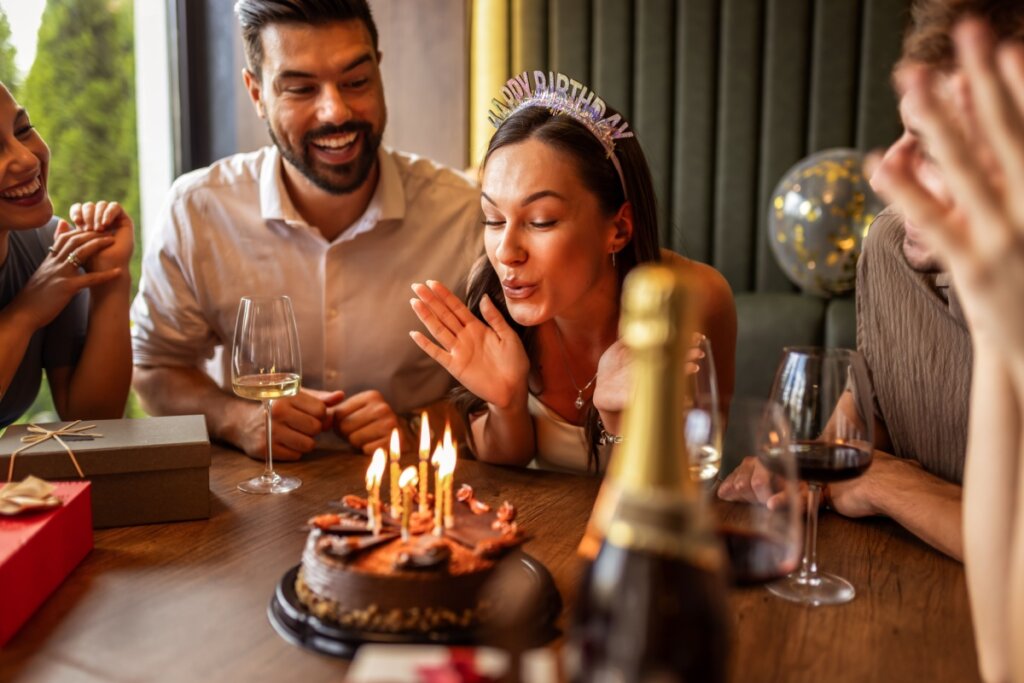 Fotografia em close de uma celebração de aniversário em uma mesa de restaurante. Uma mulher usando uma tiara que diz "Happy Birthday" está prestes a assoprar as velas acesas de um bolo de chocolate. Ela está cercada por três amigos que sorriem e observam com entusiasmo. Sobre a mesa de madeira, há presentes embrulhados, taças de vinho e uma garrafa de espumante em primeiro plano, fora de foco.