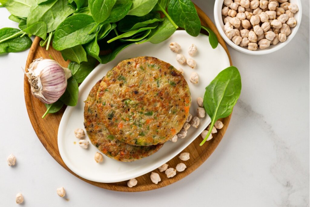 Dois hambúrgueres vegetais de grão-de-bico dourados e com cenoura e espinafre, empilhados em um prato branco oval. O prato está sobre uma tábua de madeira redonda, decorado com grãos de bico secos, uma cabeça de alho e folhas frescas de espinafre ao fundo.
