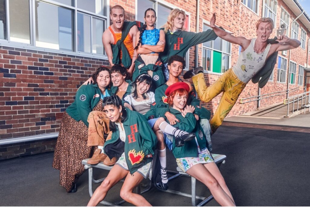 Foto promocional da série "Heartbreak High" com um grupo diversificado de cerca de dez jovens posando em frente a um prédio de tijolos vermelhos da escola. Eles vestem uniformes escolares estilizados, jaquetas verdes com a sigla "HH" e roupas coloridas e casuais. O clima é vibrante e descontraído, com alguns estudantes sorrindo e outros fazendo poses expressivas.