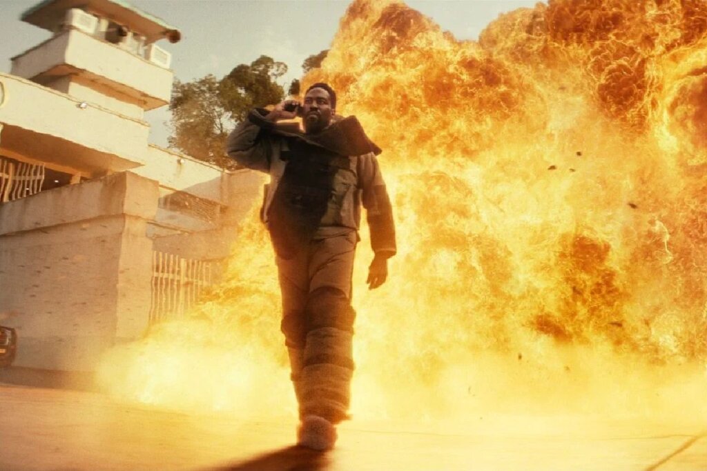 Cena de ação cinematográfica em plano médio. No centro, o ator Yahya Abdul-Mateen II, interpretando um personagem com colete tático e uma mochila pesada nas costas, caminha em direção ao espectador. Ele está falando ao celular com a mão direita e sua expressão é séria. Às suas costas, uma imensa e detalhada explosão de fogo e fumaça laranja-avermelhada consome o fundo. O cenário parece ser uma rua pavimentada diante de um complexo industrial ou prisional de concreto, com uma torre de observação visível à esquerda, sob uma luz diurna difusa.