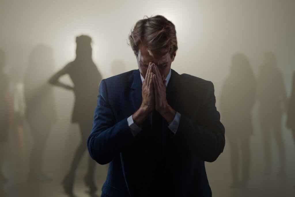 Um homem de terno azul escuro está em primeiro plano, com a cabeça baixa e as mãos unidas cobrindo o rosto, em um gesto que expressa profunda angústia, tristeza ou oração. A iluminação é dramática, deixando o homem parcialmente em sombras. Ao fundo, envoltas em uma névoa clara e desfocada, aparecem silhuetas de várias pessoas que parecem observar ou ignorar a cena. A composição visual enfatiza sentimentos de isolamento, rejeição e vulnerabilidade social.