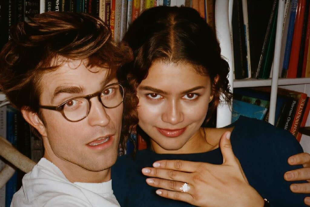 Close-up dos atores Robert Pattinson e Zendaya posando juntos. Robert, à esquerda, usa óculos de armação fina e redonda e uma camiseta branca, com uma expressão de surpresa. Zendaya, à direita, sorri levemente e posiciona a mão sobre o peito, exibindo um anel de destaque no dedo anelar. O fundo mostra uma estante repleta de livros.