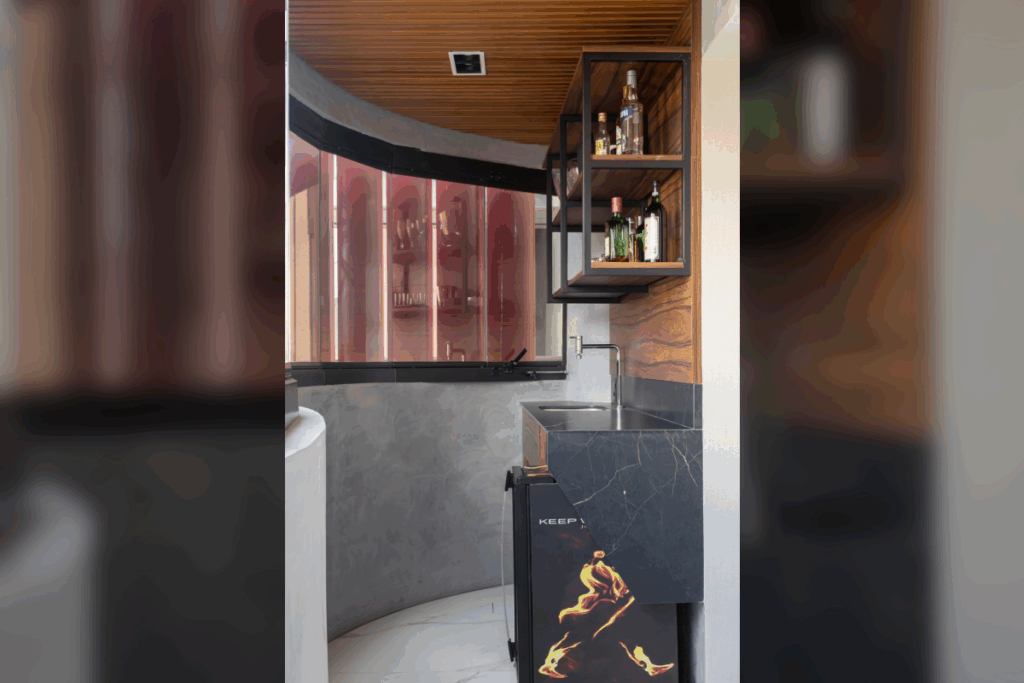 Esta imagem mostra um painel de madeira rica com veios horizontais cobrindo a parede de fundo e uma parte da parede lateral em um canto de bar moderno. Prateleiras de metal preto e madeira são montadas sobre o painel de madeira na parede lateral. O teto também é revestido por um painel de madeira ripado, criando uma continuidade visual no canto curvo que também apresenta uma parede de concreto aparente e um frigobar preto com design de chamas.