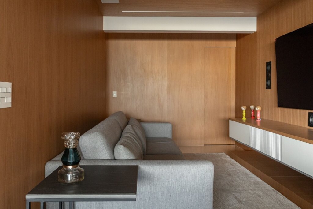 Esta imagem de uma sala de estar compacta apresenta painéis de madeira cobrindo todas as paredes visíveis. O teto também é revestido por um forro de madeira ripado, criando uma atmosfera envolvente e acolhedora. Um grande painel de madeira na parede de fundo apresenta uma porta mimetizada (camuflada) com um puxador sutil. Um painel de TV preto e um rack flutuante de madeira e branco são montados sobre o painel de madeira na parede lateral.