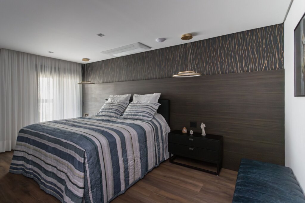 Esta imagem de um quarto moderno exibe um piso de madeira de tábuas largas. Uma parede lateral, no lado direito da imagem, é coberta por um grande painel de madeira com portas de armário embutidas e espelhadas. A parede atrás da cama apresenta um painel de cabeceira de madeira na cor terracota, que se estende verticalmente sobre uma parede com padrão de papel de parede.