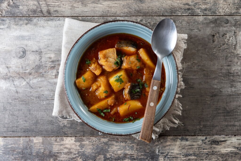 Vista superior de uma tigela de cerâmica azul contendo peixe ensopado com pedaços de batata em um molho denso e avermelhado. O prato está finalizado com ervas frescas picadas. A tigela repousa sobre um guardanapo de tecido bege, que está sobre uma mesa de madeira rústica e envelhecida. Uma colher com cabo de madeira está posicionada ao lado direito da tigela.