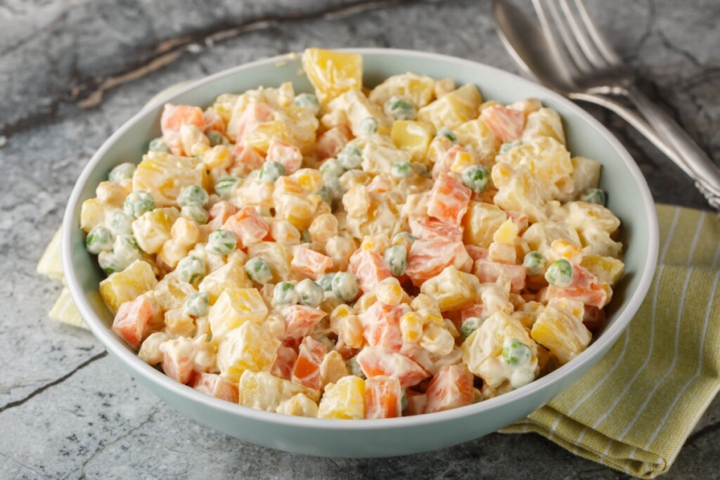 Uma tigela redonda de cor azul-clara repleta de salada de maionese. A mistura contém cubos de batata cozida, pedaços de cenoura laranja, ervilhas verdes e grãos de milho amarelo, todos envolvidos em maionese. A tigela está posicionada sobre uma mesa cinza com textura de mármore, ao lado de um guardanapo de tecido listrado e talheres de metal ao fundo.