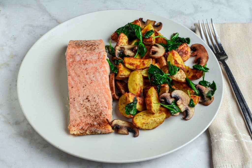 Filé de salmão grelhado servido com batatas rústicas douradas, cogumelos e espinafre em prato branco