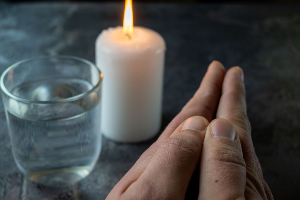 Close-up em ângulo diagonal mostrando mãos unidas em posição de oração ou reflexão no primeiro plano. Ao fundo, levemente desfocados, estão uma vela branca cilíndrica acesa, com a chama brilhante, e um copo de vidro transparente com água. O conjunto de elementos está sobre uma superfície de pedra escura e rústica, sob uma iluminação suave que destaca o contraste entre a luz da vela e as sombras.
