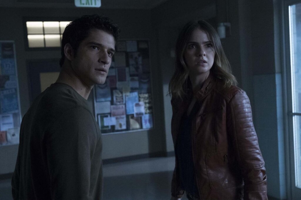 Fotografia de uma cena da série Teen Wolf. Em um ambiente interno mal iluminado que parece um corredor de escola, Scott McCall (Tyler Posey) à esquerda e Malia Tate (Shelley Hennig) à direita olham para algo fora do quadro com expressões de preocupação e alerta. Scott usa uma blusa verde escura e Malia veste uma jaqueta de couro marrom. Ao fundo, vê-se um mural com cartazes e uma placa de saída iluminada.