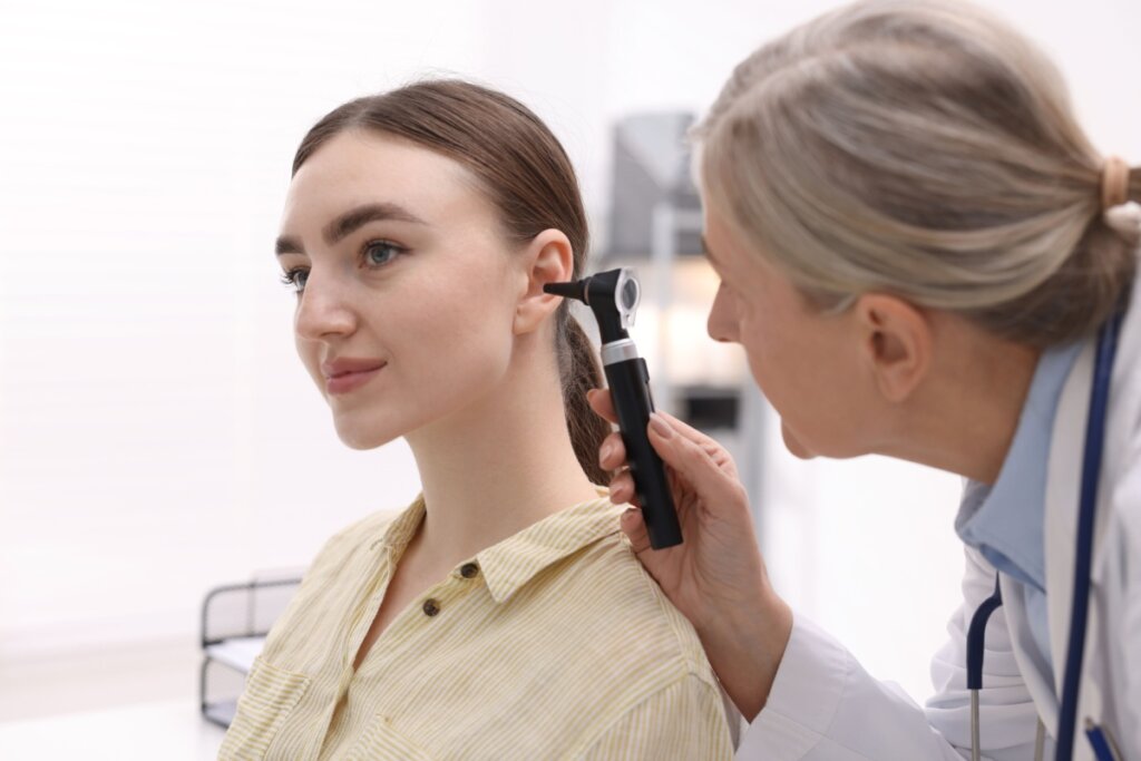 Fotografia em ambiente clínico onde uma médica examina o ouvido de uma paciente jovem com um otoscópio. A médica, uma mulher de cabelos grisalhos presa em um rabo de cavalo, está de perfil à direita. A paciente, à esquerda, veste uma camisa listrada amarela e branca e olha calmamente para a frente. O fundo é claro e levemente desfocado.