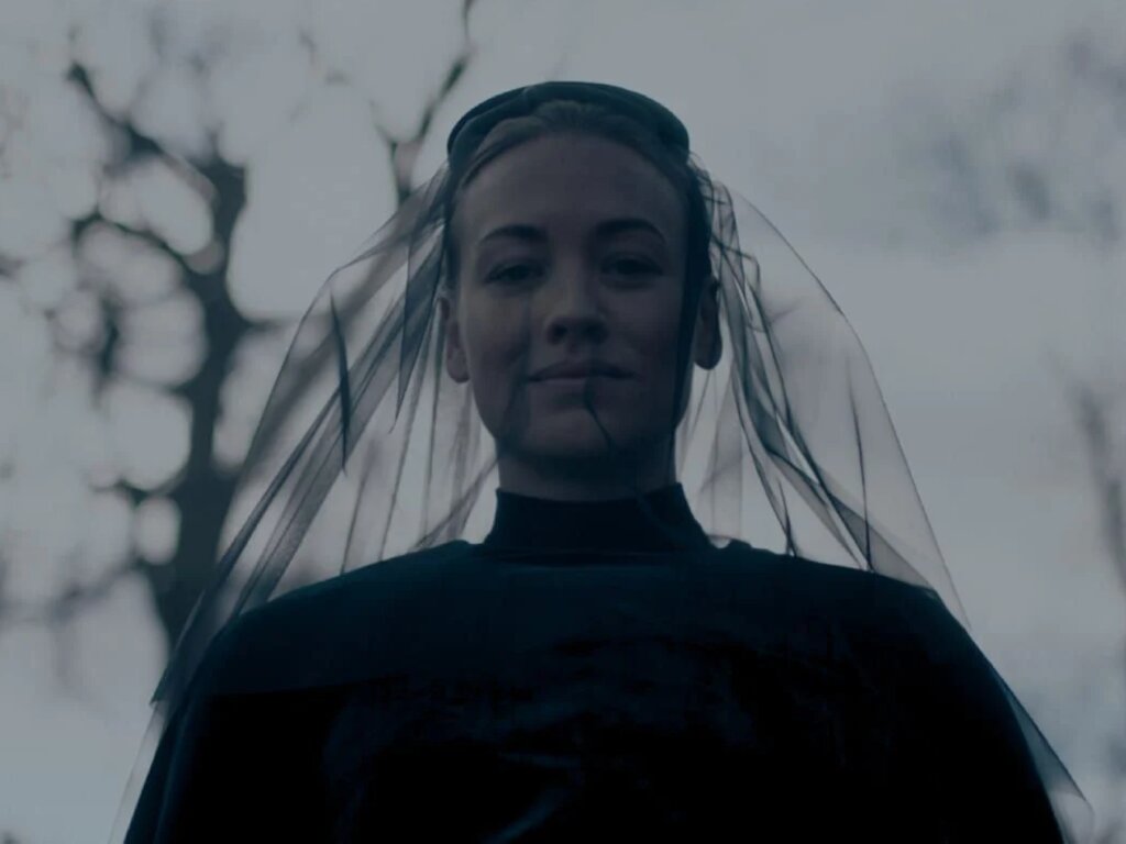 Close-up frontal de Serena Joy (Yvonne Strahovski) da série "The Handmaid’s Tale". Ela está centralizada na imagem, olhando diretamente para a câmera com uma expressão serena e um leve sorriso. Veste roupas pretas de luto e um véu de tule preto que cobre parcialmente seu rosto e desce pelos ombros. O fundo está desfocado, mostrando galhos secos de árvores sob um céu nublado e acinzentado, criando uma atmosfera fria e solene.