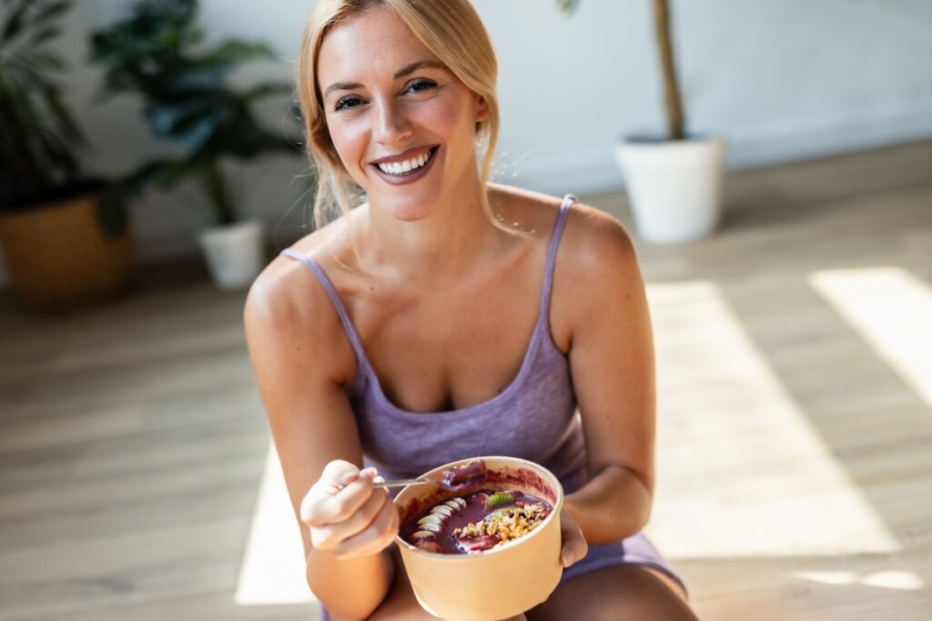 Mulher loira com conjunto roxo de academia sorrindo e segurando bowl com açaí e banana