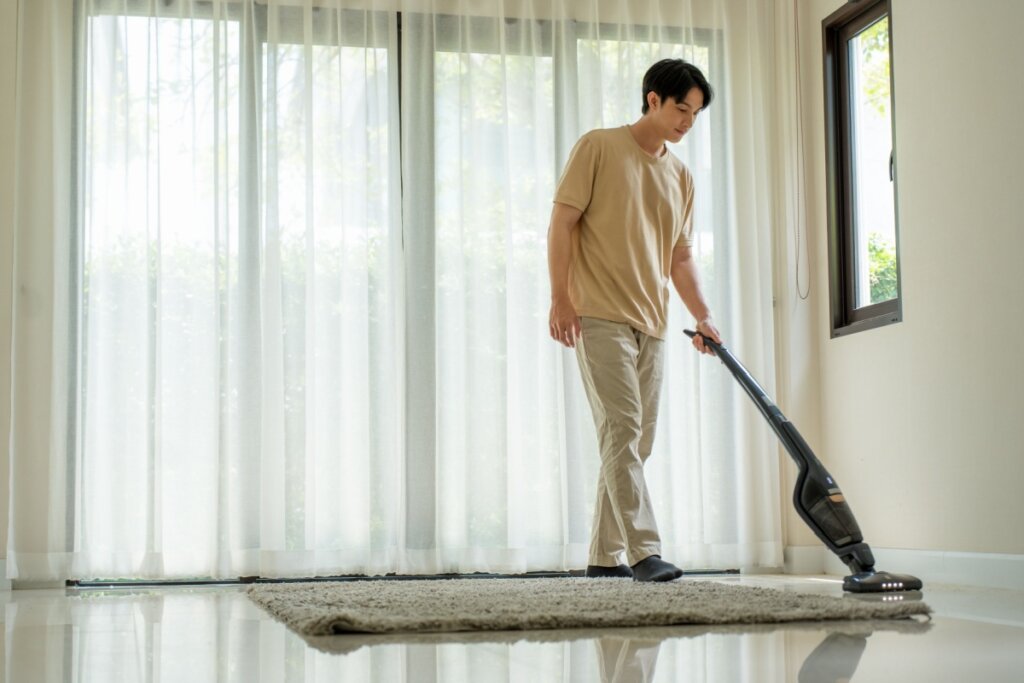 Homem passando aspirador em um tapete bege em frente a uma janela com cortina branca 