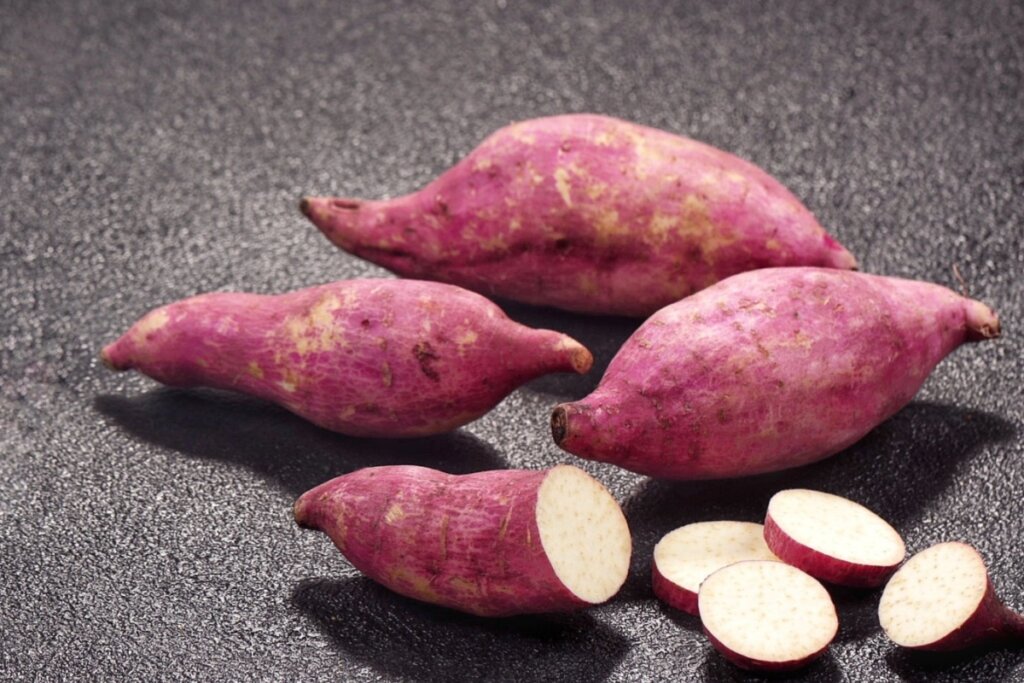 Três batatas doces inteiras e uma fatiada até a metade em uma bancada escura.