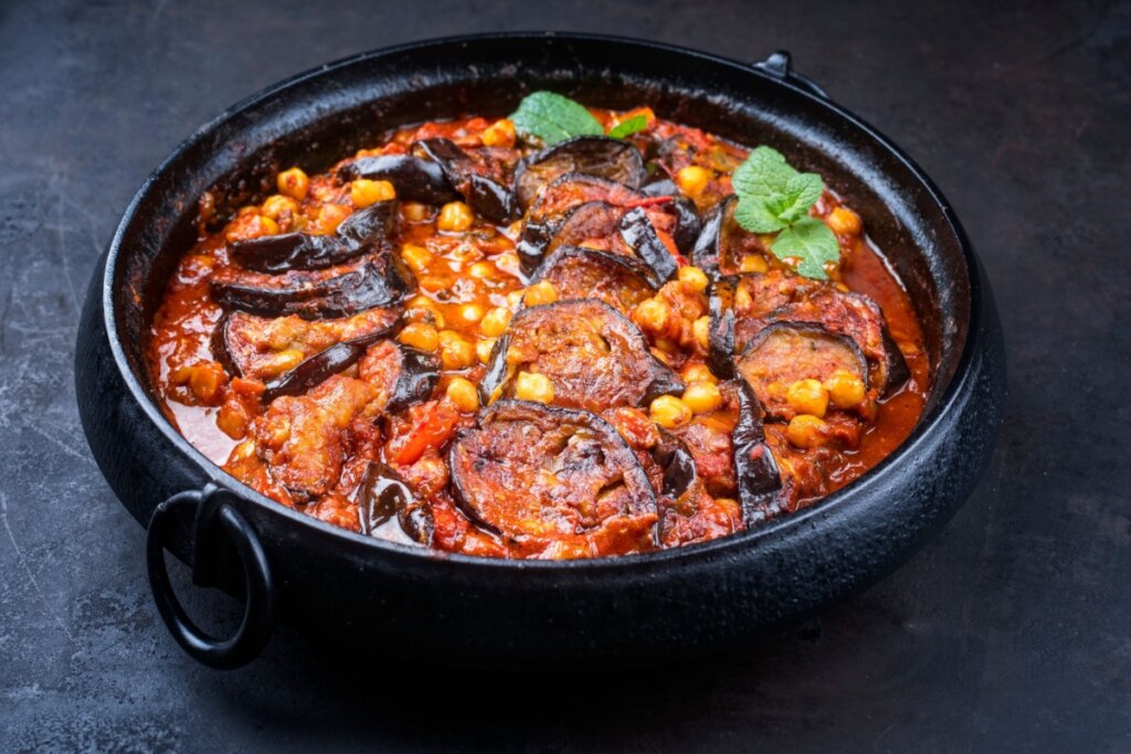 A imagem mostra um prato servido em uma panela escura com um ensopado de berinjela em rodelas, grão-de-bico e molho vermelho. O prato está decorado com folhas verdes por cima, sugerindo uma refeição quente e bem temperada.