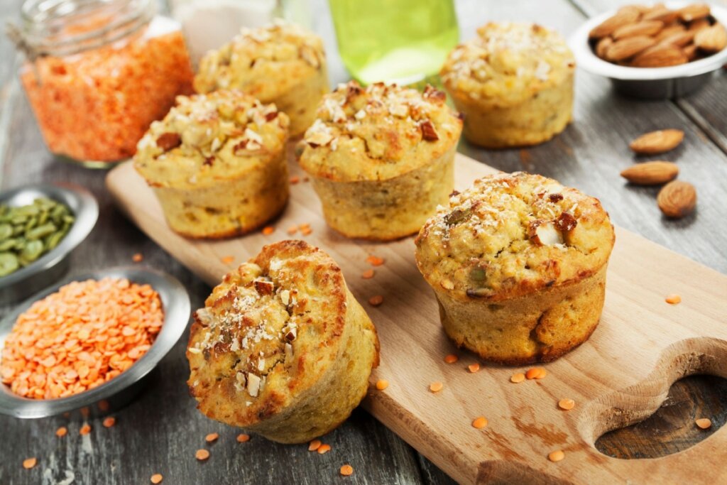 A imagem mostra vários bolinhos assados colocados sobre uma tábua de madeira. Ao redor deles aparecem ingredientes como lentilhas vermelhas, sementes e amêndoas, sugerindo que os bolinhos foram preparados com esses alimentos.