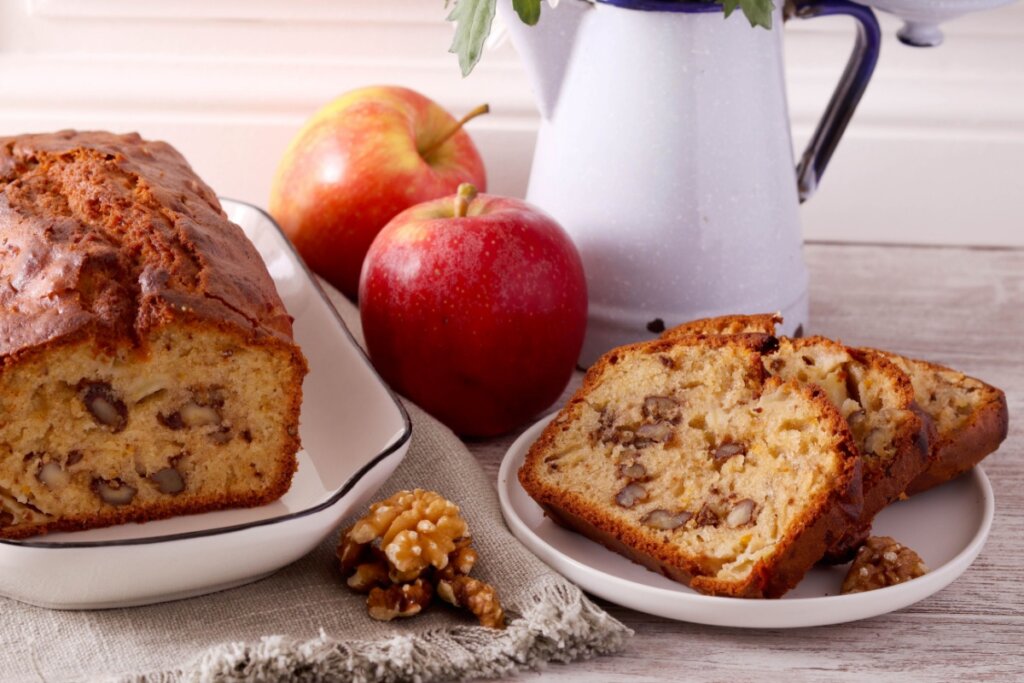 prato branco com duas fatias de bolo de maçã e nozes. ao lado prato maior com o bolo inteiro. duas maçãs vermelhas ao meio e bule de café