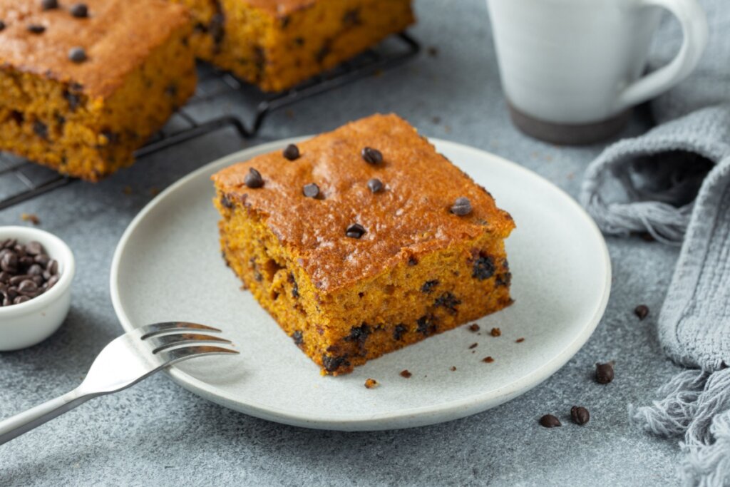 Fatia de bolo de abóbora com gotas de chocolate servida em prato branco em cima de bancada de pedra