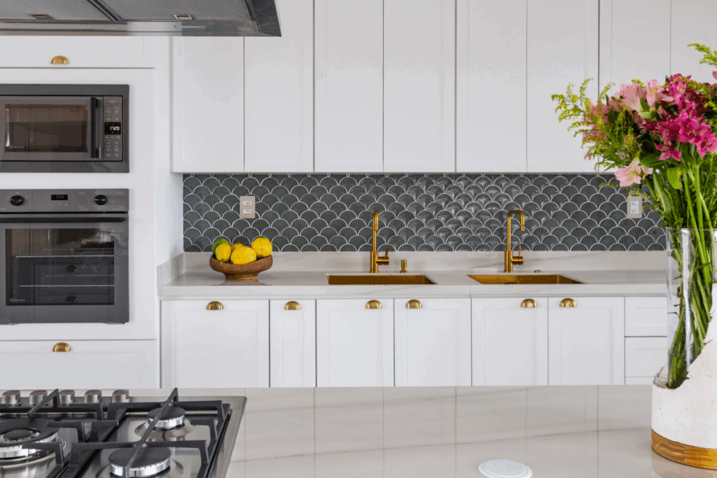 Cozinha branca com dupla cuba dourada e revestimento em escama verde compondo o fundo da bancada