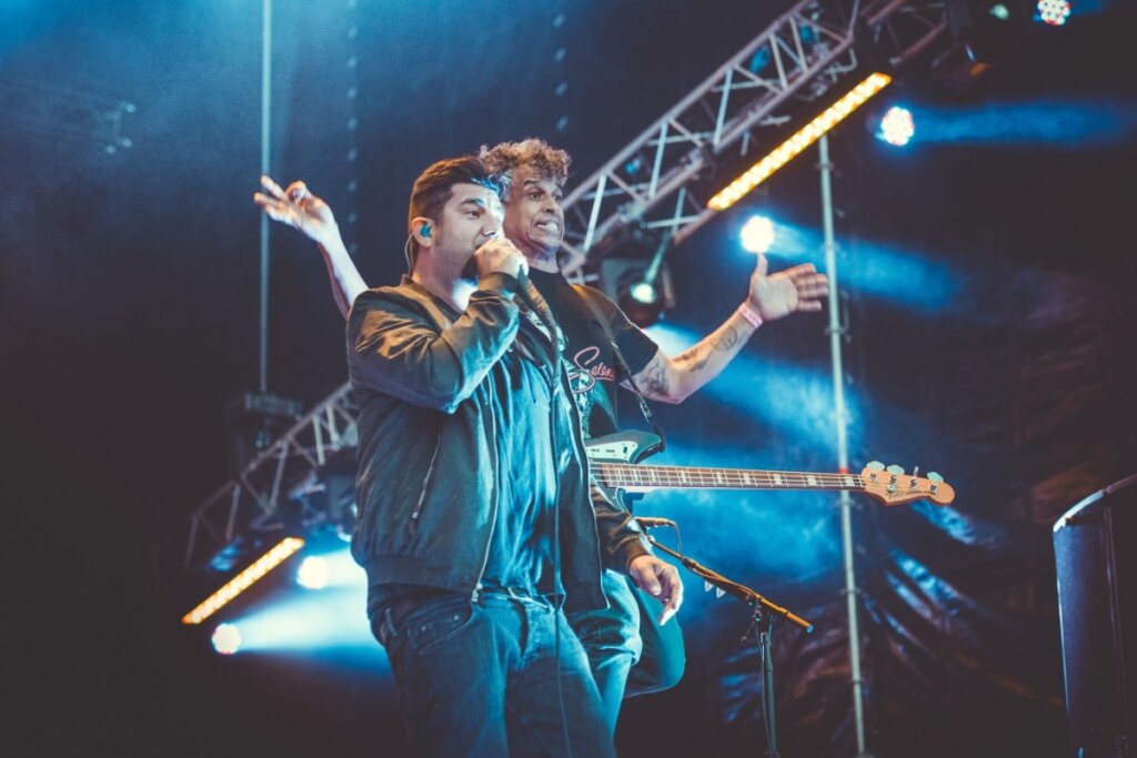 Homem com cabelo curto e cavanhaque usando calça preta e jaqueta preta cantando e atrás um homem com os dois braços abertos e com baixo em cima de palco