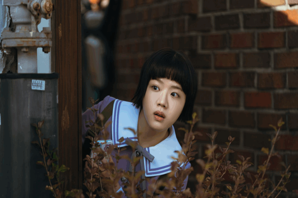 Imagem de uma jovem com o cabelo preto curto, usando uniforme escolar escondida atrás de árvore