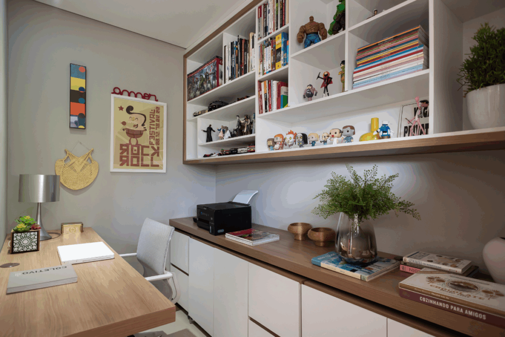 Escritório com móveis em tons de bege e madeira, com mesa de computador com tampo de madeira claro, bancada com gavetas e impressora e itens de decoração e estante suspensa