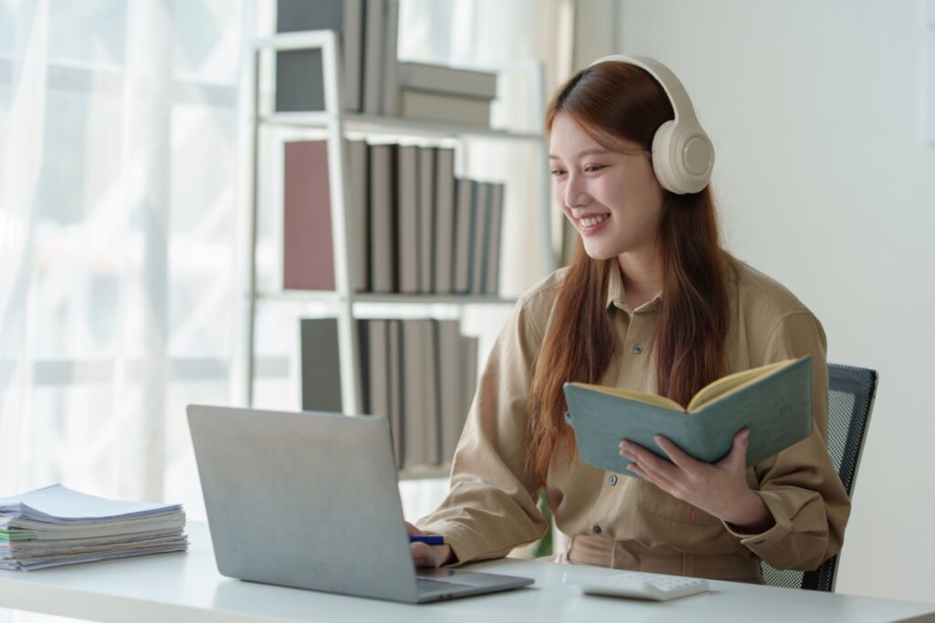 Jovem com cabelo liso solto, usando camisa de manga longa bege e com um livro na mão e com fone de ouvido e com computador a frente estudando atualidades