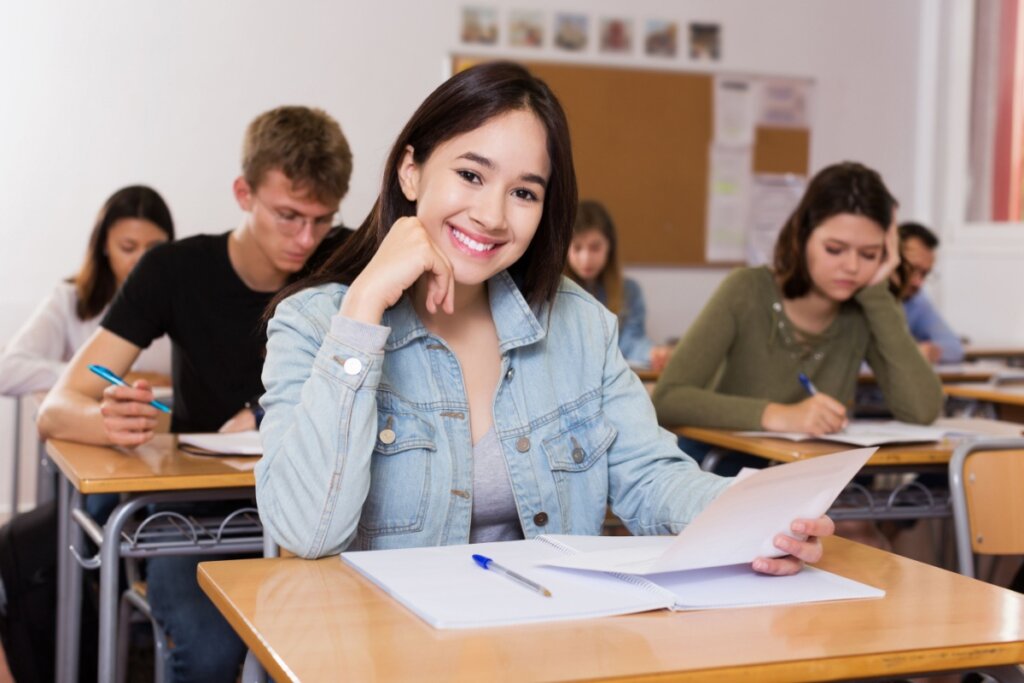 jovem de jaqueta jeans, sentada em sala de aula, com braço apoiado na mesa, sorrindo e segurando papel. apostila com caneta em cima da mesa