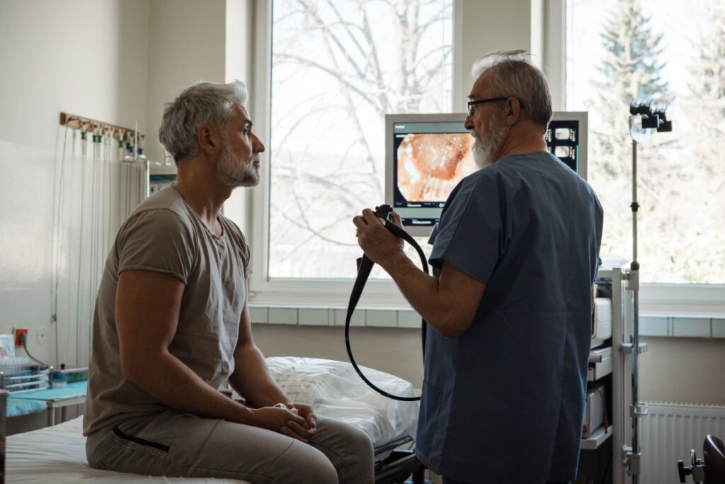 Homem sentado com médico segurando um aparelho de exame de colonoscopia na sua frente